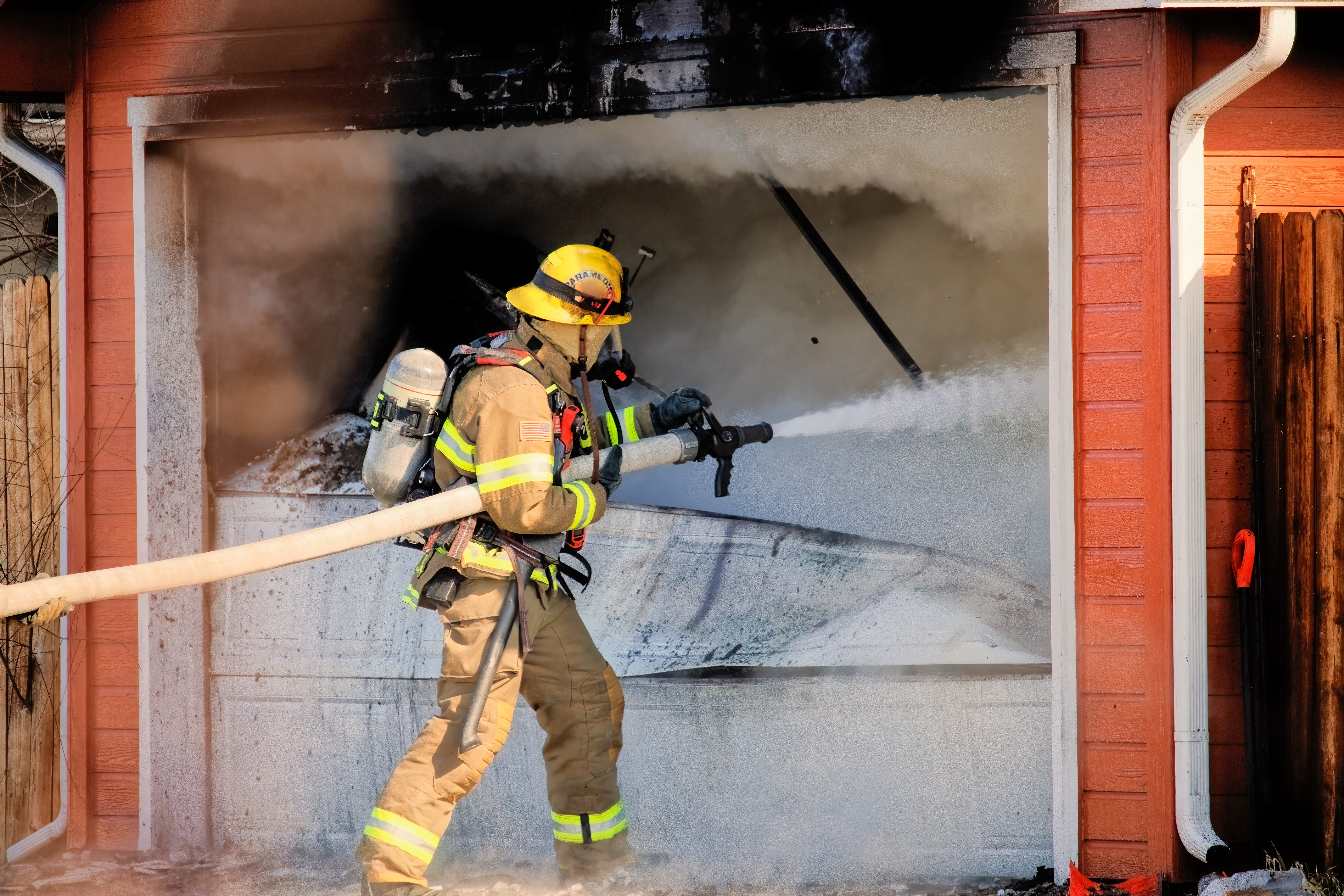 Firefighter using a hose to extinguish a residential house fire, showing suppression water damage risk during fire restoration.